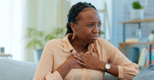 A young Black woman sitting on the couch looking distressed while putting both hands on her chest, likely due to an irregular heartbeat