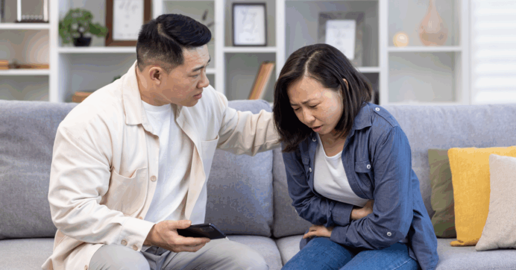 a young Asian woman, experiencing an ulcerative colitis (UC) flare-up, clutches her stomach while being comforted by her husband on a couch.