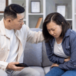a young Asian woman, experiencing an ulcerative colitis (UC) flare-up, clutches her stomach while being comforted by her husband on a couch.