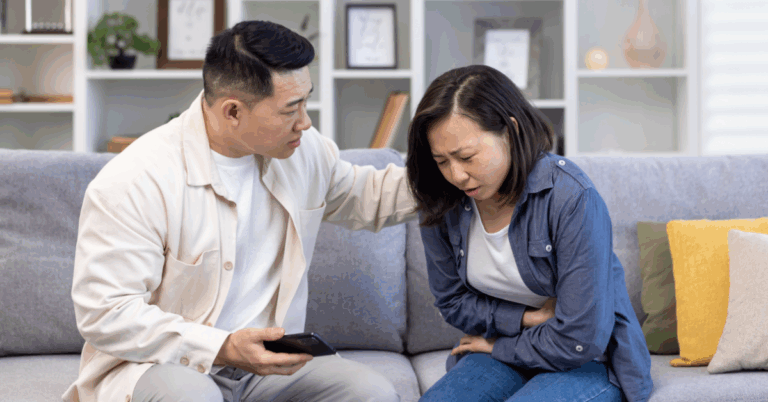 a young Asian woman, experiencing an ulcerative colitis (UC) flare-up, clutches her stomach while being comforted by her husband on a couch.