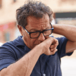 An older man, centered in his neighborhood, covers his mouth while coughing. This action suggests a link to poor air quality, a probable contributor to his lung cancer risk.