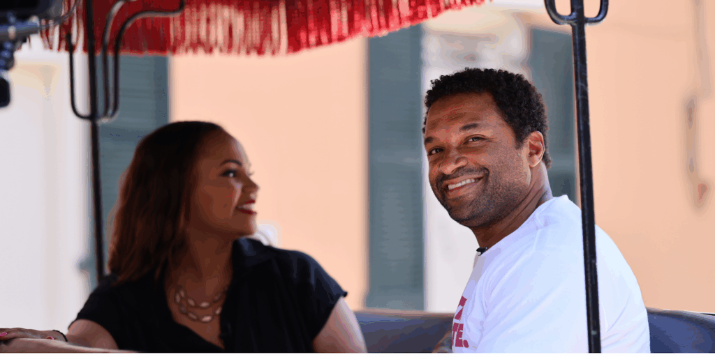 Del Smith in a white t-shirt smiles directly at the camera while sitting in the back of a brightly colored, open-air vehicle. Tiffany sitting in the vehicle, slightly out of focus in the foreground.