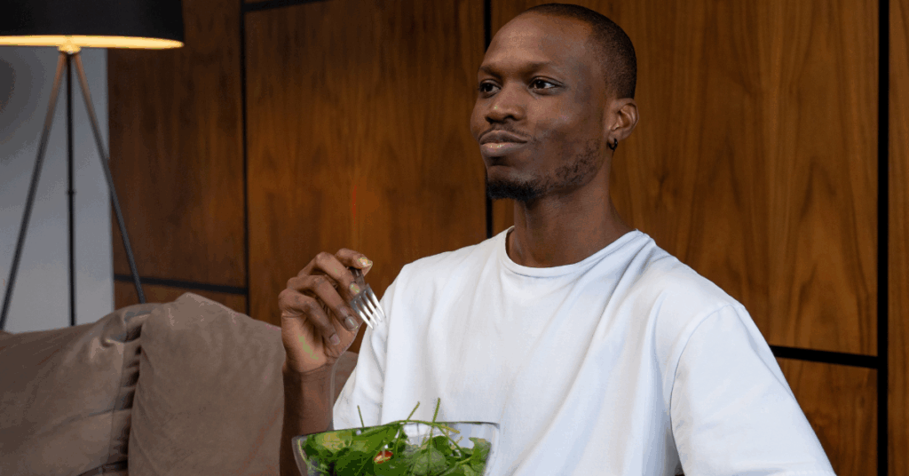 Happy young Black man eating a spinach salad sitting on the couch, which could be affecting his kidney health.