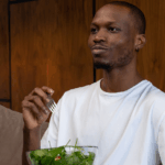 Happy young Black man eating a spinach salad sitting on the couch, which could be affecting his kidney health.