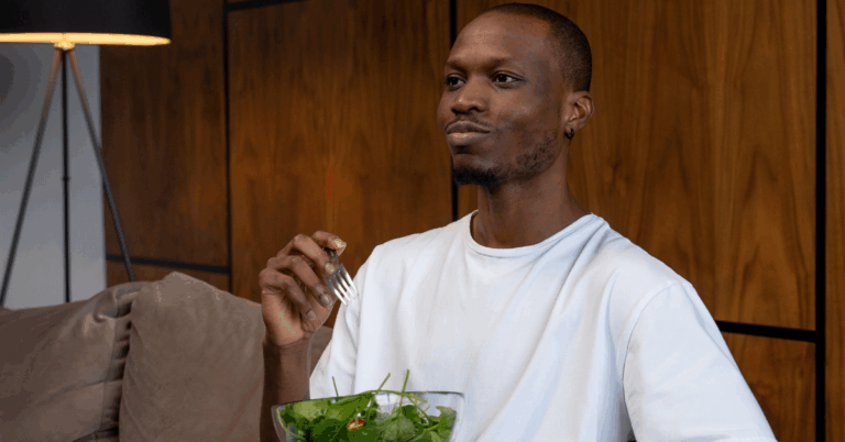 Happy young Black man eating a spinach salad sitting on the couch, which could be affecting his kidney health.