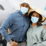 Young African American couple wearing masks and beach hats, standing in front of a plane, ready to travel
