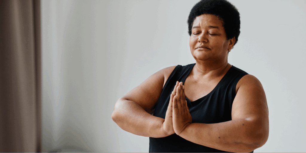 A Black woman with short black hair meditates with closed eyes and hands in a prayer position, practicing self-care and mindfulness to manage stress during her lung cancer treatment.