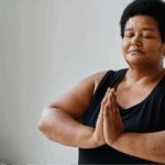 A Black woman with short black hair meditates with closed eyes and hands in a prayer position, practicing self-care and mindfulness to manage stress during her lung cancer treatment.