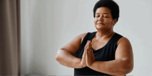 A Black woman with short black hair meditates with closed eyes and hands in a prayer position, practicing self-care and mindfulness to manage stress during her lung cancer treatment.
