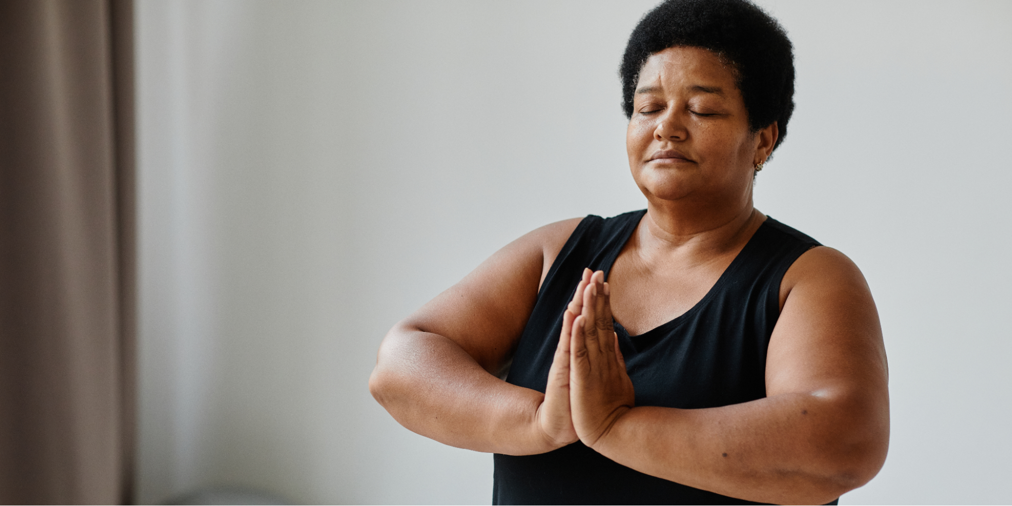 A Black woman with short black hair meditates with closed eyes and hands in a prayer position, practicing self-care and mindfulness to manage stress during her lung cancer treatment.