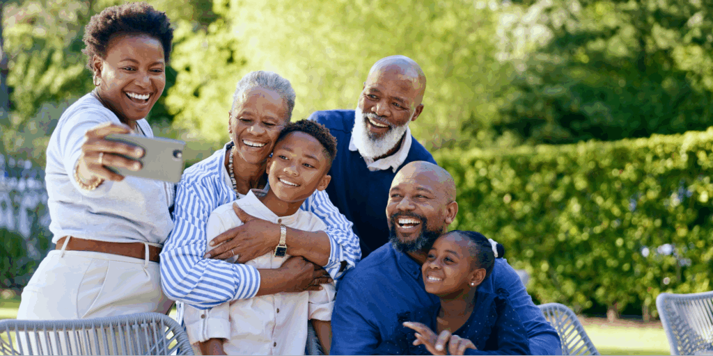 A multi-generational Black family of six, including two grandparents, two adults, and two children, smiling happily while taking a group selfie outdoors in a sunny backyard.