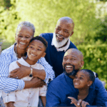 A multi-generational Black family of six, including two grandparents, two adults, and two children, smiling happily while taking a group selfie outdoors in a sunny backyard.
