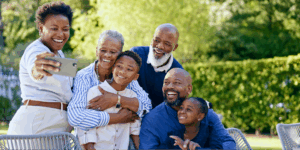 A multi-generational Black family of six, including two grandparents, two adults, and two children, smiling happily while taking a group selfie outdoors in a sunny backyard.