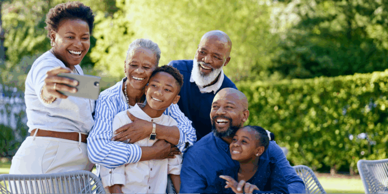 A multi-generational Black family of six, including two grandparents, two adults, and two children, smiling happily while taking a group selfie outdoors in a sunny backyard.