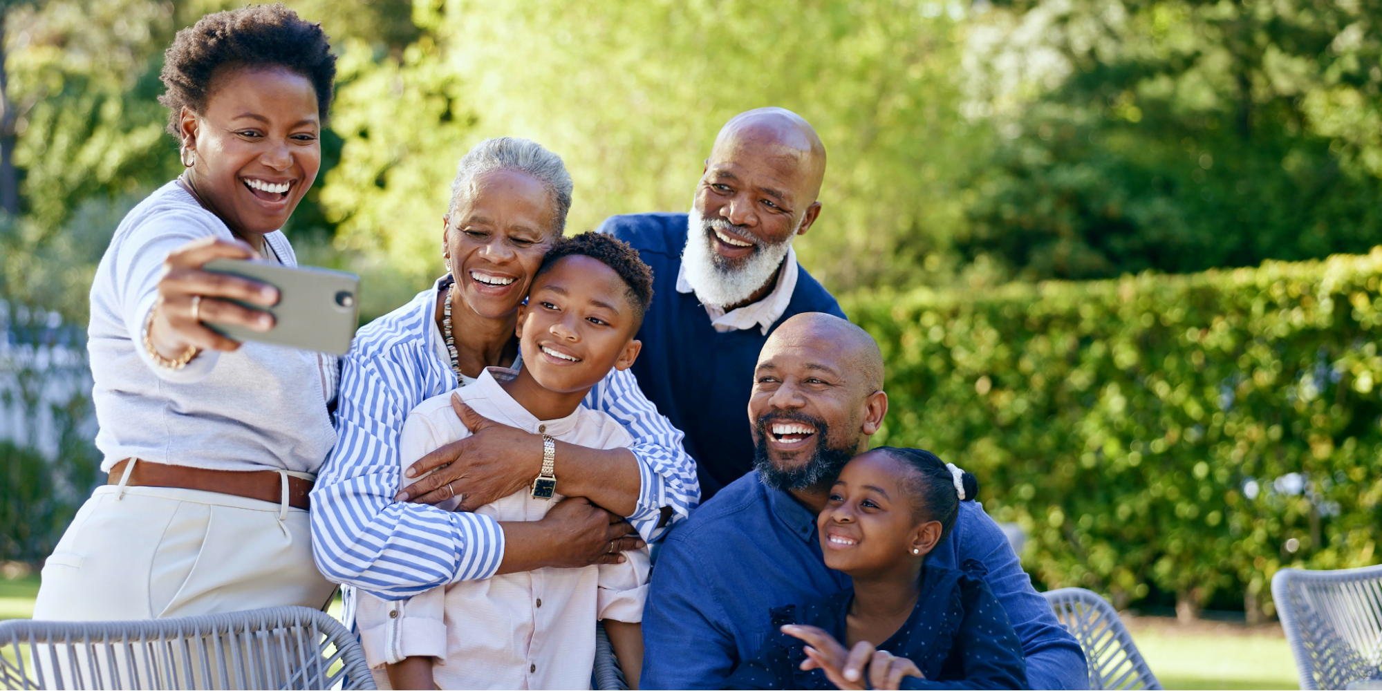 A multi-generational Black family of six, including two grandparents, two adults, and two children, smiling happily while taking a group selfie outdoors in a sunny backyard.