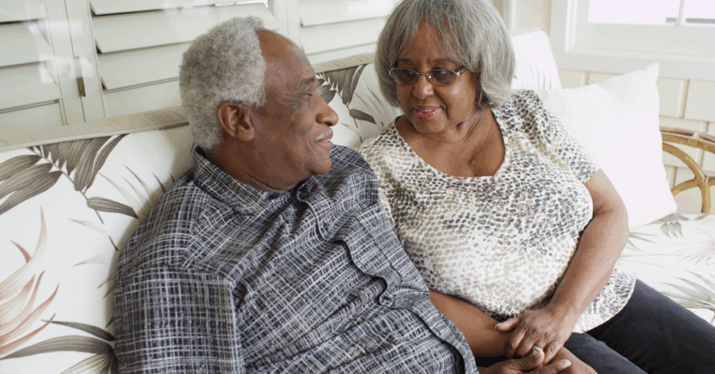 An older Black couple, sitting on the couch, conversing. The woman is holding the man’s hand as he may be showing signs of early dementia.