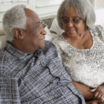 An older Black couple, sitting on the couch, conversing. The woman is holding the man’s hand as he may be showing signs of early dementia.