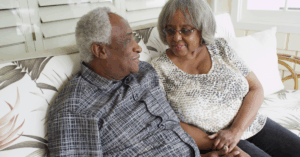 An older Black couple, sitting on the couch, conversing. The woman is holding the man’s hand as he may be showing signs of early dementia.