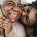 Close-up image of an older Black woman with gray hair, likely living with breast cancer, and a younger Black woman with curly hair hugging, both smiling with eyes closed.