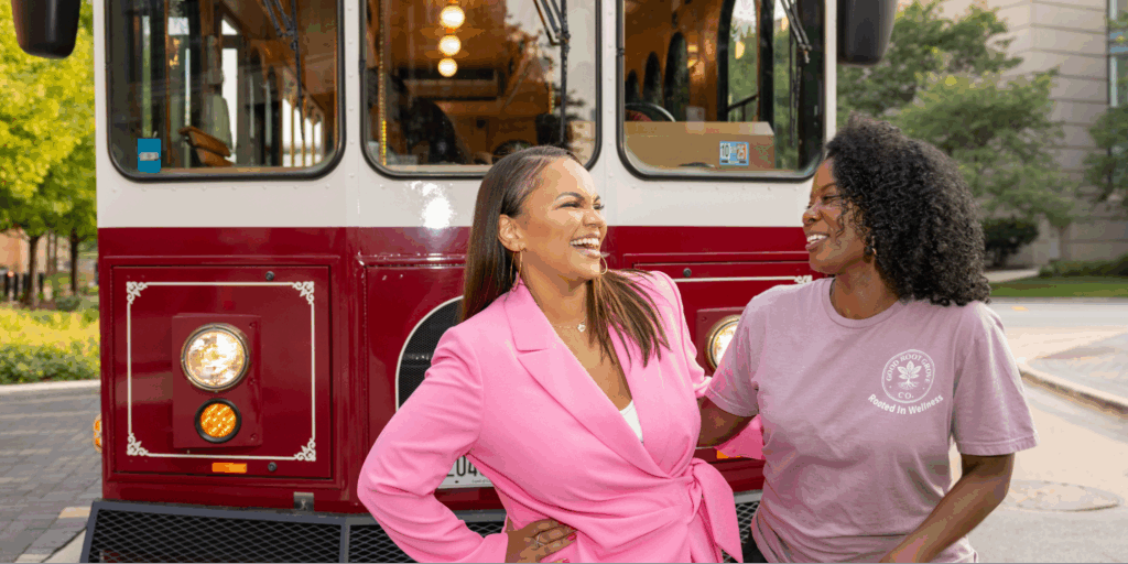 Tiffany Whitlow, Co-founder and CDO, Acclinate and Michelle Brantley-Smith, founder of The Good Root Grove Co, laughing together while standing in front of a red and white vintage trolley outdoors.