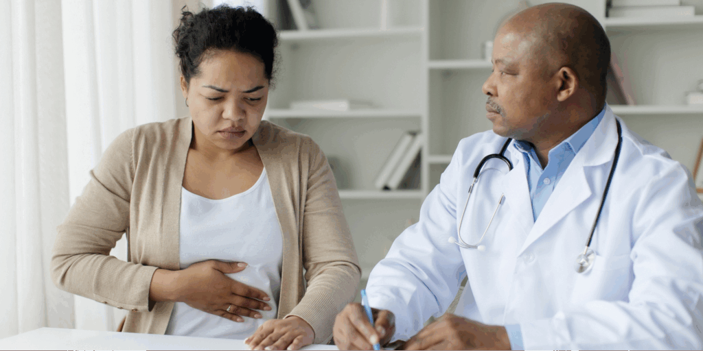 A woman expressing stomach discomfort to a doctor, illustrating the need to advocate for a colonoscopy when your pain is dismissed.