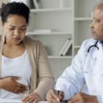 A woman expressing stomach discomfort to a doctor, illustrating the need to advocate for a colonoscopy when your pain is dismissed.