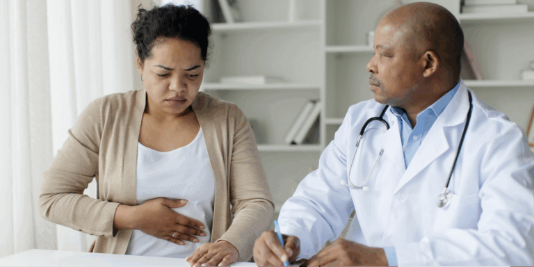 5 Black Celebrities With Lung Cancer 2 A woman expressing stomach discomfort to a doctor, illustrating the need to advocate for a colonoscopy when your pain is dismissed.