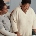 A Black female social worker helps a senior woman walk with a walker, demonstrating the personalized support available through affordable home care and adult day services.