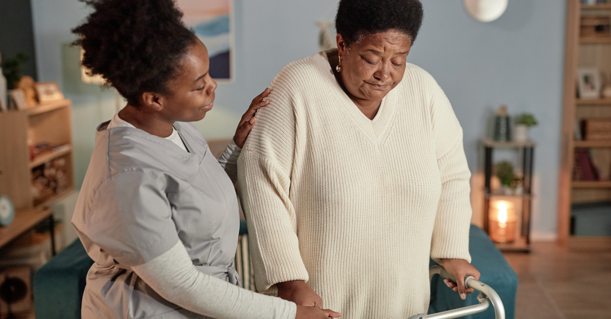 A Black female social worker helps a senior woman walk with a walker, demonstrating the personalized support available through affordable home care and adult day services.