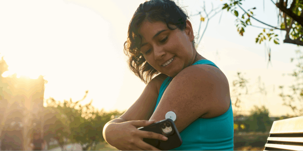 A close-up of a Hispanic woman in athletic wear managing her health by using a Continuous Glucose Monitor (CGM) attached to her arm.