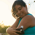 A close-up of a Hispanic woman in athletic wear managing her health by using a Continuous Glucose Monitor (CGM) attached to her arm.