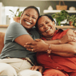 A close-up of two Black women smiling and embracing at home, highlighting the importance of shared health awareness to lower your breast cancer risk.
