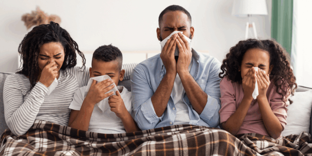 A family of four sitting on a couch under a blanket, all blowing their noses with tissues while sick with allergies or the flu.