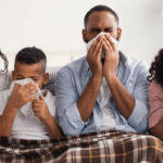 A family of four sitting on a couch under a blanket, all blowing their noses with tissues while sick with allergies or the flu.