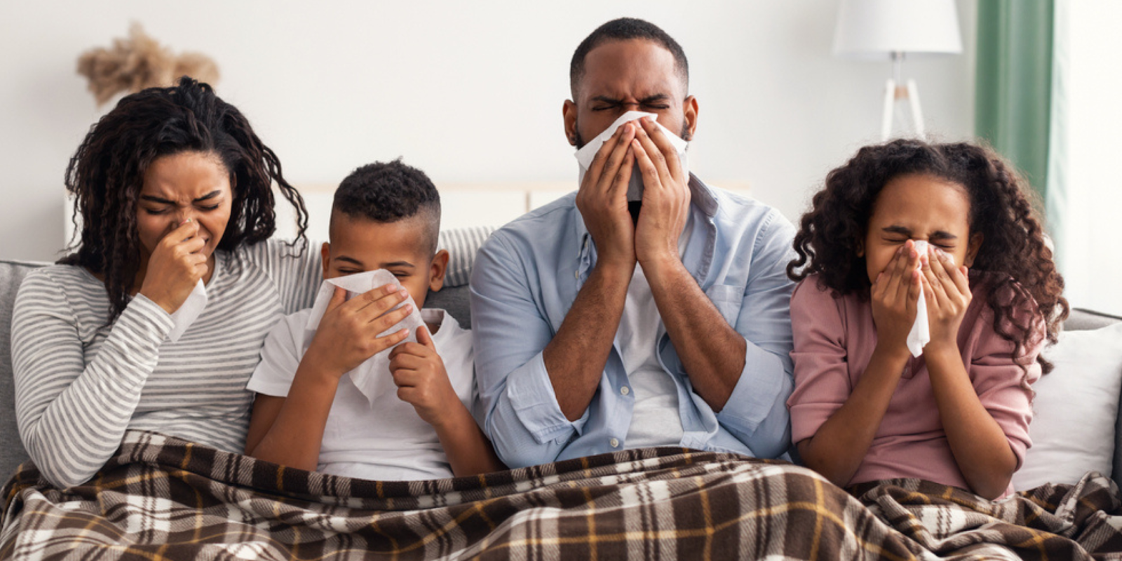 A family of four sitting on a couch under a blanket, all blowing their noses with tissues while sick with allergies or the flu.