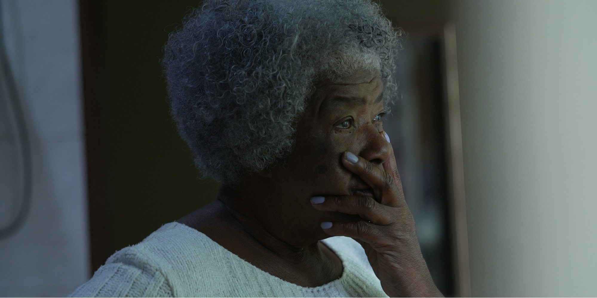 A Black elderly woman looking out a window appearing troubled by memory loss caused by normal aging or dementia.