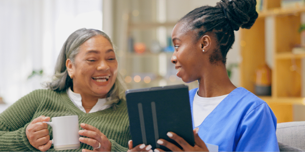 A young Black female caregiver showing a tablet to a Hispanic senior woman teaching her to find and apply for breast cancer financial assistance programs.