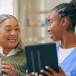 A young Black female caregiver showing a tablet to a Hispanic senior woman teaching her to find and apply for breast cancer financial assistance programs.