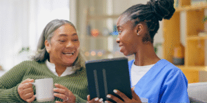 A young Black female caregiver showing a tablet to a Hispanic senior woman teaching her to find and apply for breast cancer financial assistance programs.