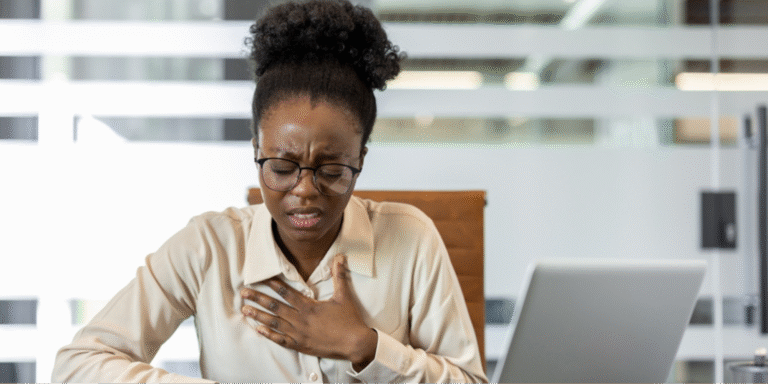 A Black woman in an office setting experiencing chest discomfort, representing the uncertainty of whether symptoms are related to stress or heart disease.
