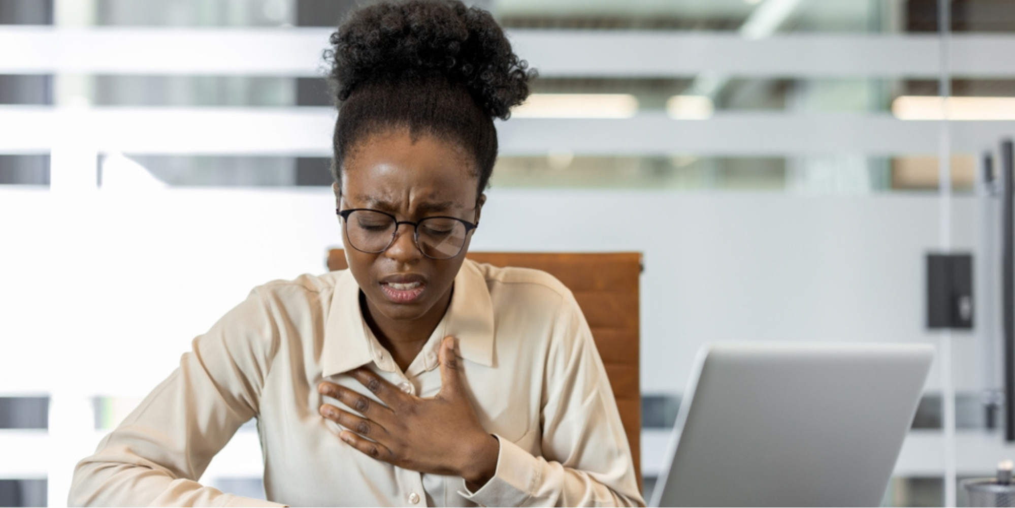 A Black woman in an office setting experiencing chest discomfort, representing the uncertainty of whether symptoms are related to stress or heart disease.