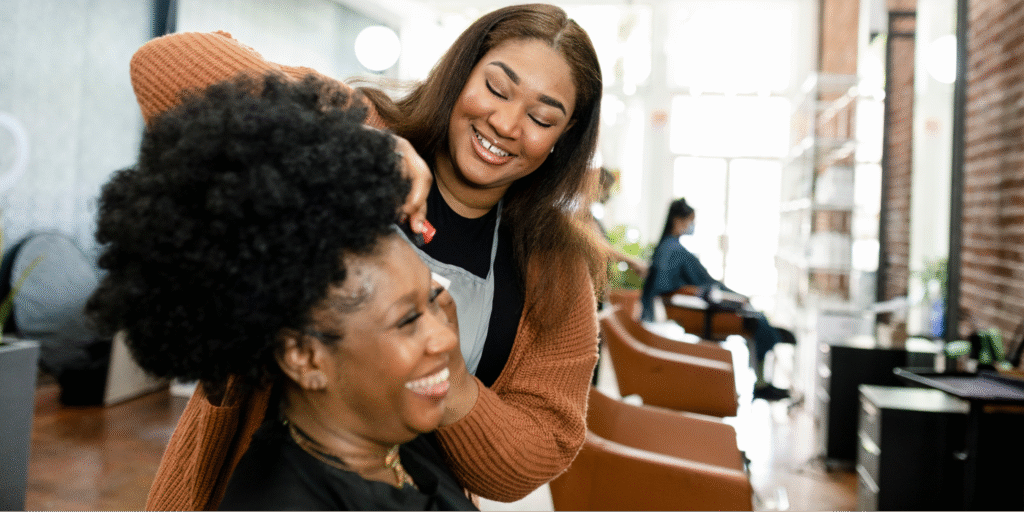 A joyful interaction at a hair salon between a Black hairstylist and a Black woman, highlighting the importance of specialized cancer wigs.