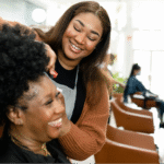 A joyful interaction at a hair salon between a Black hairstylist and a Black woman, highlighting the importance of specialized cancer wigs.