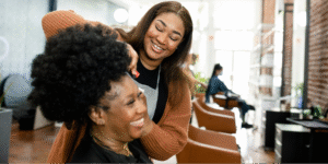 A joyful interaction at a hair salon between a Black hairstylist and a Black woman, highlighting the importance of specialized cancer wigs.