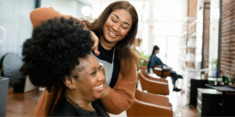 A joyful interaction at a hair salon between a Black hairstylist and a Black woman, highlighting the importance of specialized cancer wigs.