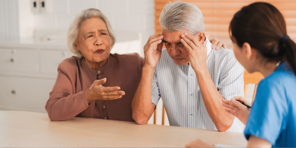 An elderly Asian man holding his head in distress while his wife talks to a nurse, illustrating the importance of knowing what to ask your doctor about brain health questions and memory screenings.