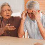 An elderly Asian man holding his head in distress while his wife talks to a nurse, illustrating the importance of knowing what to ask your doctor about brain health questions and memory screenings.