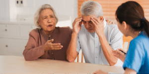 An elderly Asian man holding his head in distress while his wife talks to a nurse, illustrating the importance of knowing what to ask your doctor about brain health questions and memory screenings.