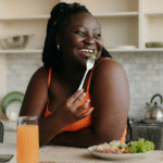 A Black woman in an orange tanktop, sitting on a chair next to the kitchen counter, and eating low fiber high protein meals