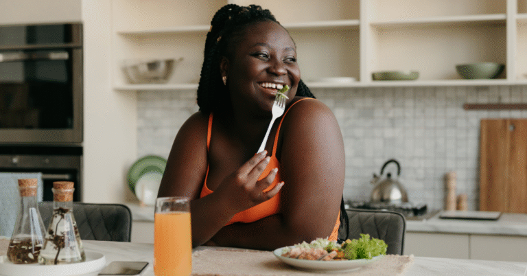 A Black woman in an orange tanktop, sitting on a chair next to the kitchen counter, and eating low fiber high protein meals